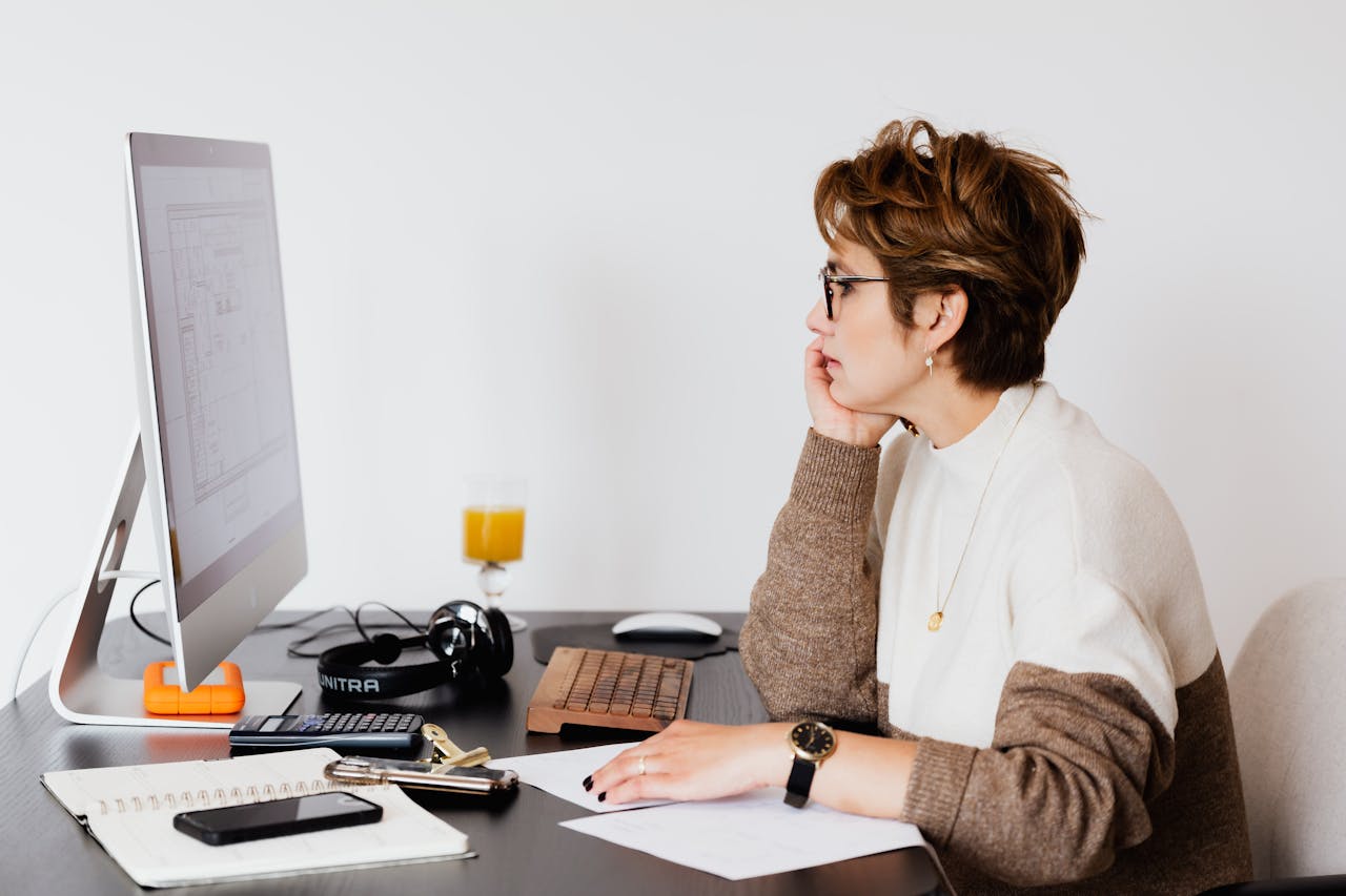 Offerings Side view of elegant female architect in glasses sitting at desk with modern PC and studying plan of building on computer screen